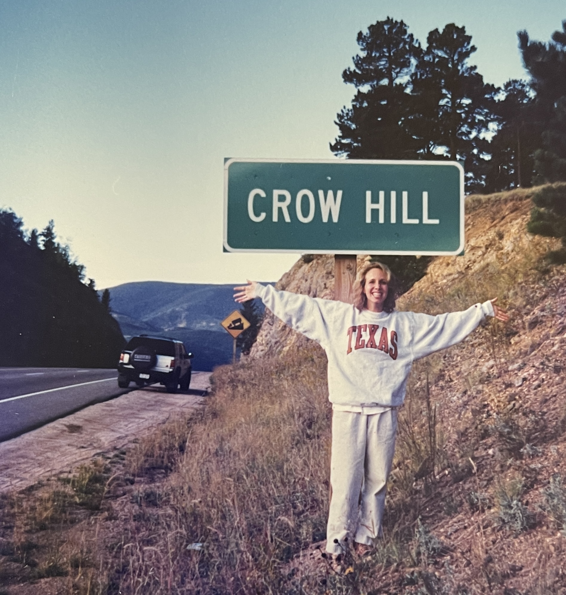 Allison standing on the side of hwy 285 in colorado with her arms open wide. Just behind her is a sign that says CROW HILL and beyond that her 1995 Isuzu rodeo is parked on the edge of the road.