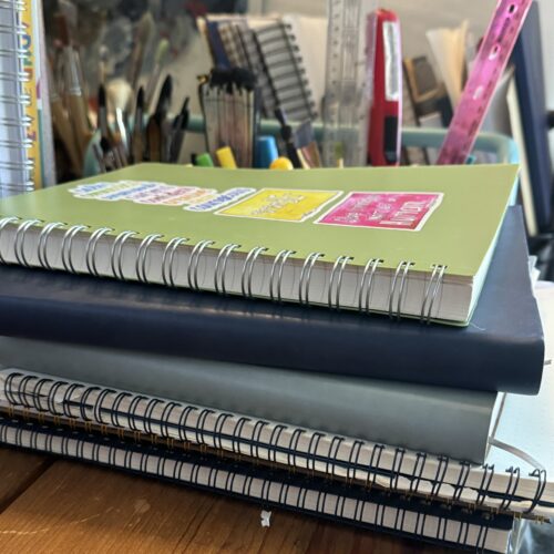 journals2024 A stack of spiral and leather bound journals on allison's desk.